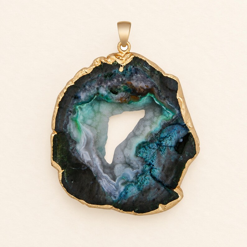 May include: A pendant featuring a raw, irregular slice of agate stone with a central hollow opening. The stone displays deep shades of blue, green, and black, with a textured, crystalline interior. The outer edge is encased in a gold-toned metal.