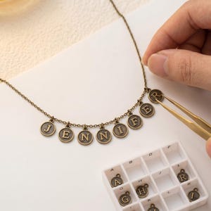 May include: A vintage-style necklace spelling "JENNIFER" with bronze-toned letter charms. The necklace is being assembled with tweezers. A tray of spare letter charms is also visible.