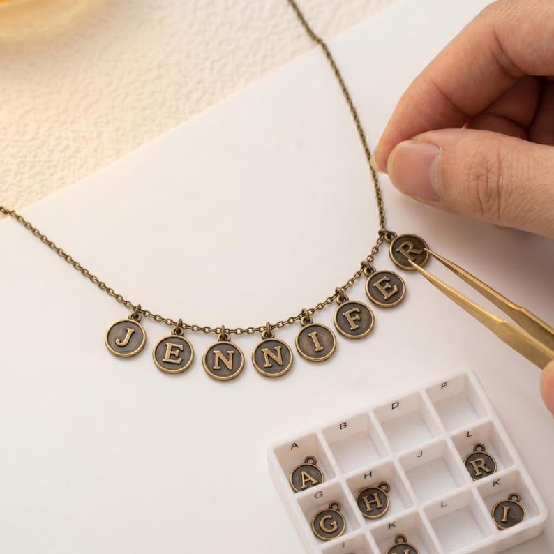 May include: A vintage-style necklace spelling "JENNIFER" with bronze-toned letter charms. The necklace is being assembled with tweezers. A tray of spare letter charms is also visible.