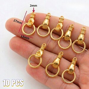 May include: Ten gold-colored hand-shaped charms with a ring. Each charm is approximately 1 inch long, 0.5 inches wide, and 0.08 inches thick. The charms are designed to be used for jewelry making or crafting.