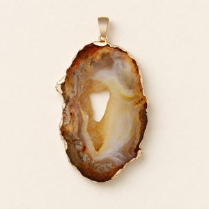 May include: A pendant featuring an irregular, translucent agate slice with concentric bands of brown, cream, and amber tones. The stone has a jagged, gold-toned metallic edge and a small, gold-toned bail at the top for attachment to a chain.