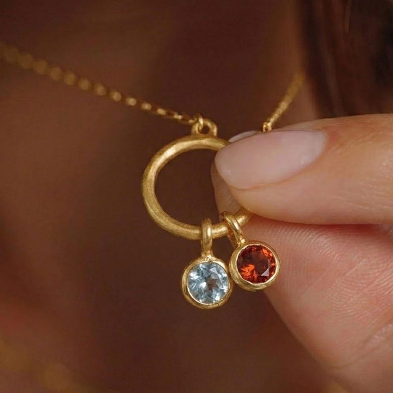 May include: A gold-colored necklace with a delicate chain and a circular pendant. The pendant holds two gemstone charms: a light blue stone and a red-orange stone. The necklace is being held by a person.