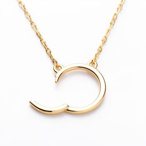 May include: A gold-colored necklace with a unique open-circle pendant. The pendant has a curved design with a small, hinged section that opens. The necklace chain is also gold-colored and delicate, creating a minimalist jewelry piece.