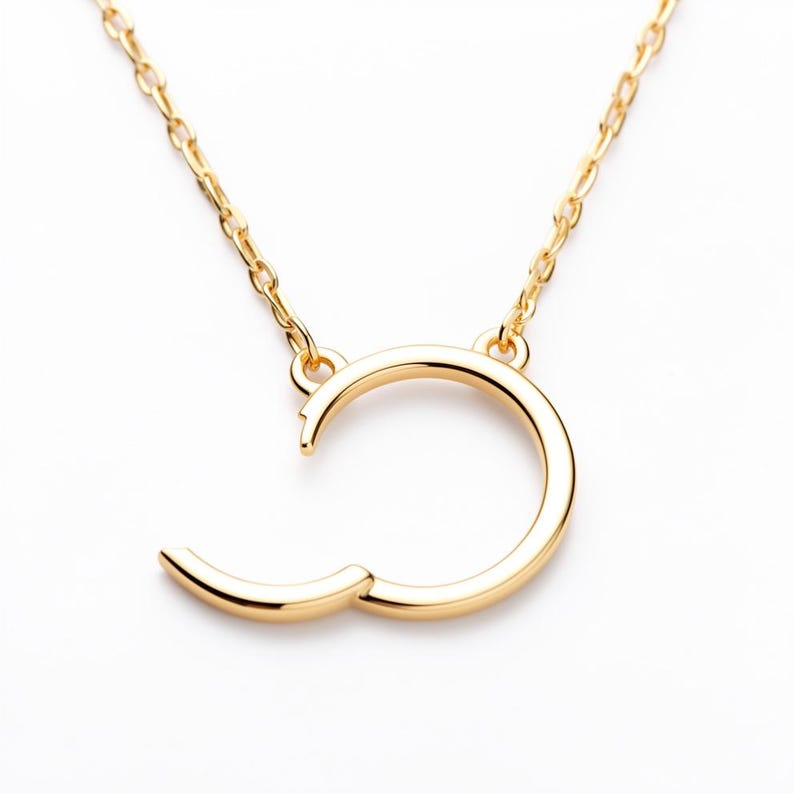 May include: A gold-colored necklace with a unique open-circle pendant. The pendant has a curved design with a small, hinged section that opens. The necklace chain is also gold-colored and delicate, creating a minimalist jewelry piece.
