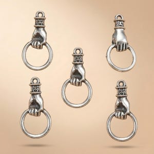 May include: Five silver-toned metal charms, each shaped like a hand grasping a circular ring. The design features a detailed cuff at the top with a small loop for attachment. The metal has a polished finish with darkened recessed areas.