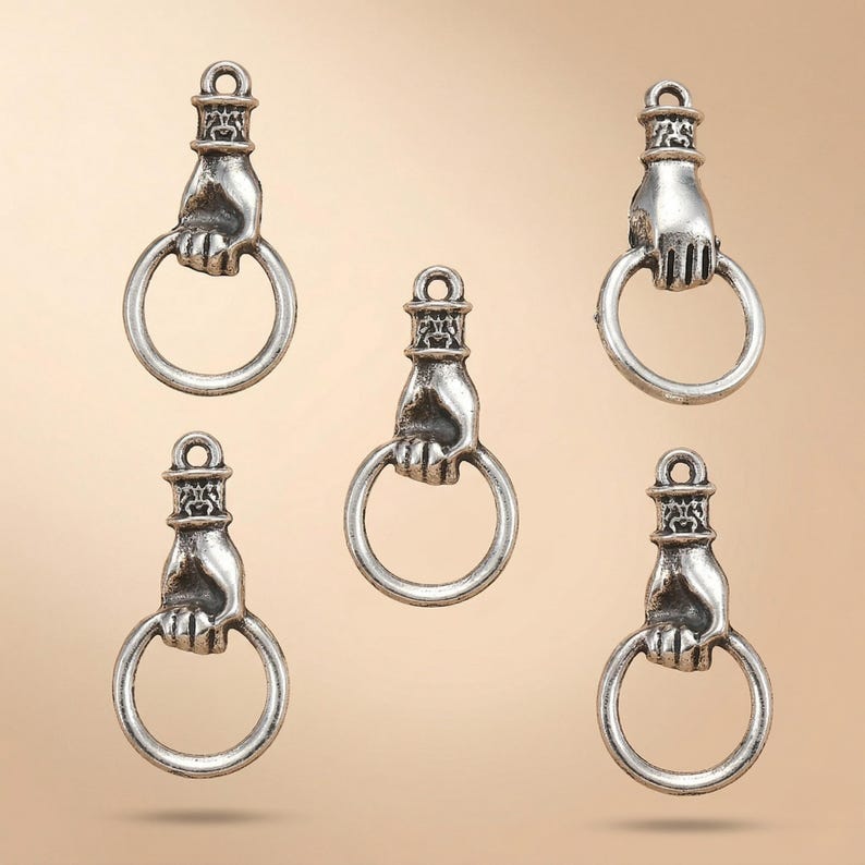 May include: Five silver-toned metal charms, each shaped like a hand grasping a circular ring. The design features a detailed cuff at the top with a small loop for attachment. The metal has a polished finish with darkened recessed areas.