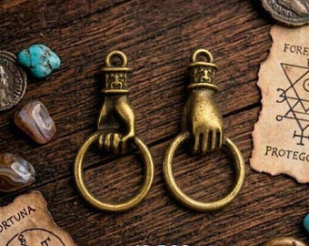 May include: Two antique brass-colored hand-shaped charms holding a ring, with the text "X10 PCS" at the bottom. The charms are on a wooden surface, surrounded by small stones and antique coins.