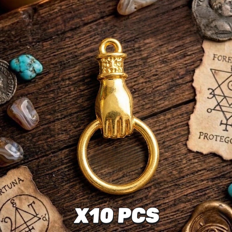 May include: A gold-colored charm in the shape of a hand holding a ring. The charm is on a wooden surface with other small objects. The text "X10 PCS" is at the bottom of the image.