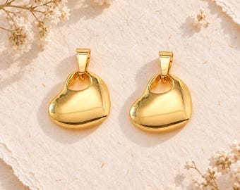 Könnte beinhalten: Ein Paar goldfarbene herzförmige Anhänger mit einer glatten, polierten Oberfläche. Jeder Anhänger hat ein abgerundetes, asymmetrisches Herzdesign mit einer kleinen integrierten Öse an der Oberseite zur Befestigung und zeigt eine reflektierende metallische Oberfläche.