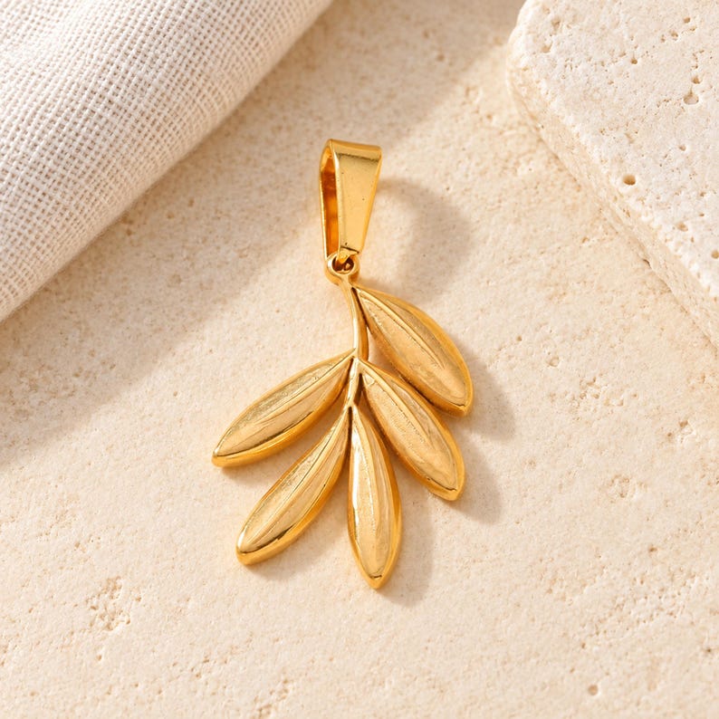 May include: A gold-toned metal pendant shaped like a small branch with five smooth, elongated leaves. The pendant features a polished finish and a sturdy, integrated bail at the top for attachment to a chain or cord, showcasing a botanical design.