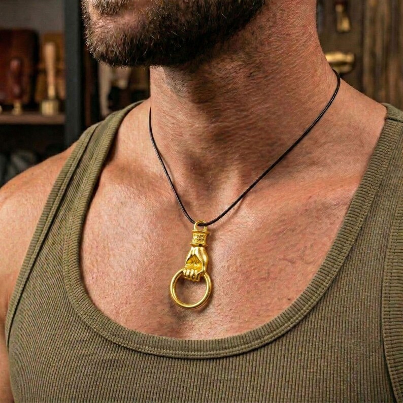 Puede incluir: Un colgante de color dorado con forma de mano que sostiene un anillo, suspendido de un fino cord&oacute;n negro. El colgante presenta un pu&ntilde;o detallado que sujeta un aro circular, creando un accesorio met&aacute;lico distintivo para el cuello.