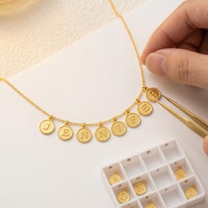 May include: A gold necklace spelling out "JENNIFER" with individual letter charms. The letters are on round, disc-shaped pendants. A hand uses tweezers to place a letter. A small white tray with more letter charms is in the foreground.