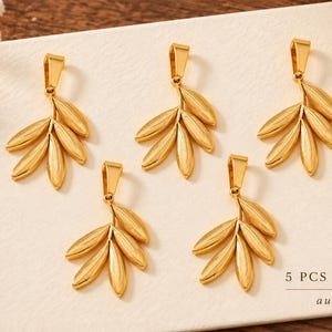 May include: Five gold-toned metal leaf-shaped pendants, each featuring a bail at the top. The pendants are arranged in a cluster, and the text 5 PCS SHOWN and the word aureza are printed in the bottom right corner of the display surface.