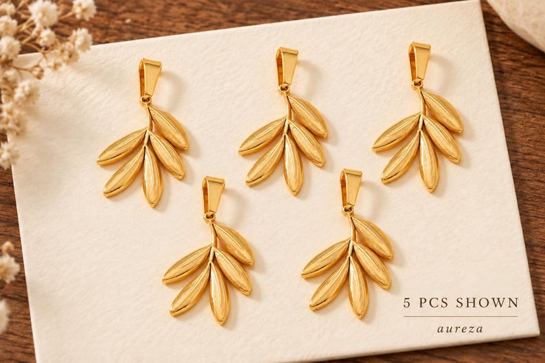 May include: Five gold-toned metal leaf-shaped pendants, each featuring a bail at the top. The pendants are arranged in a cluster, and the text 5 PCS SHOWN and the word aureza are printed in the bottom right corner of the display surface.