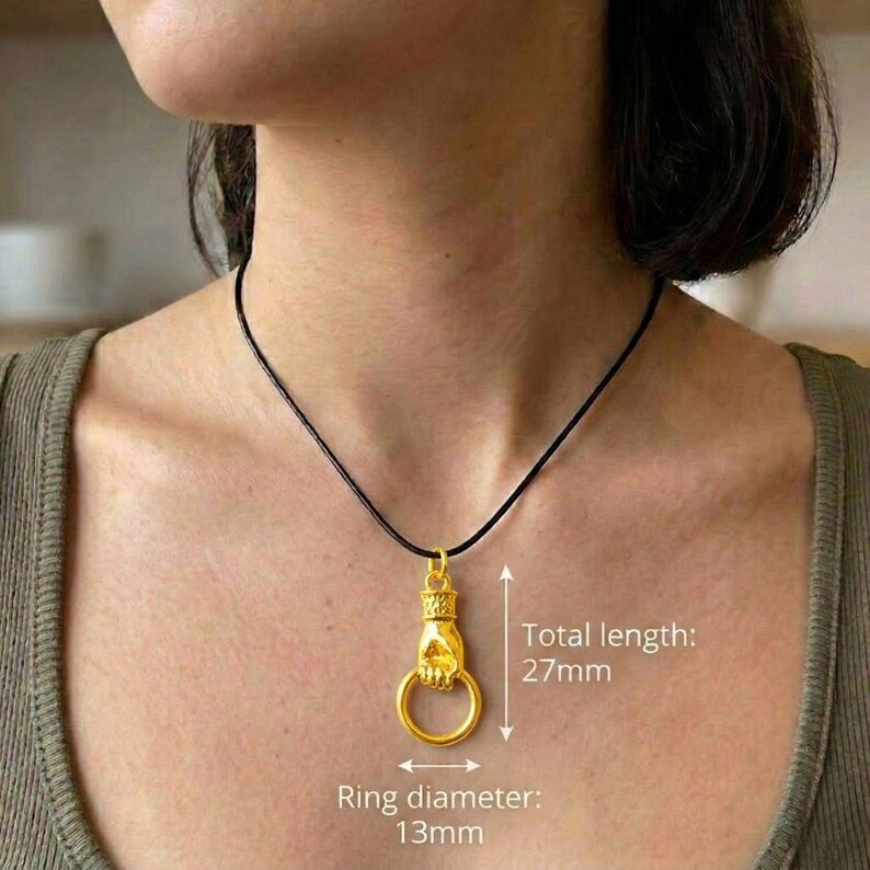 Puede incluir: Collar con colgante dorado que representa una mano sosteniendo un anillo. El colgante cuelga de un cord&oacute;n negro. El di&aacute;metro del anillo es de 13 mm, y la longitud total del colgante es de 27 mm.