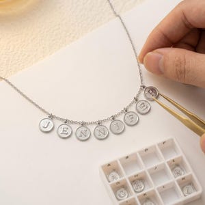 May include: A silver necklace spelling out "JENNI" with individual letter charms. The letter "R" is being added with tweezers. A small tray with spare letter charms is in the foreground.