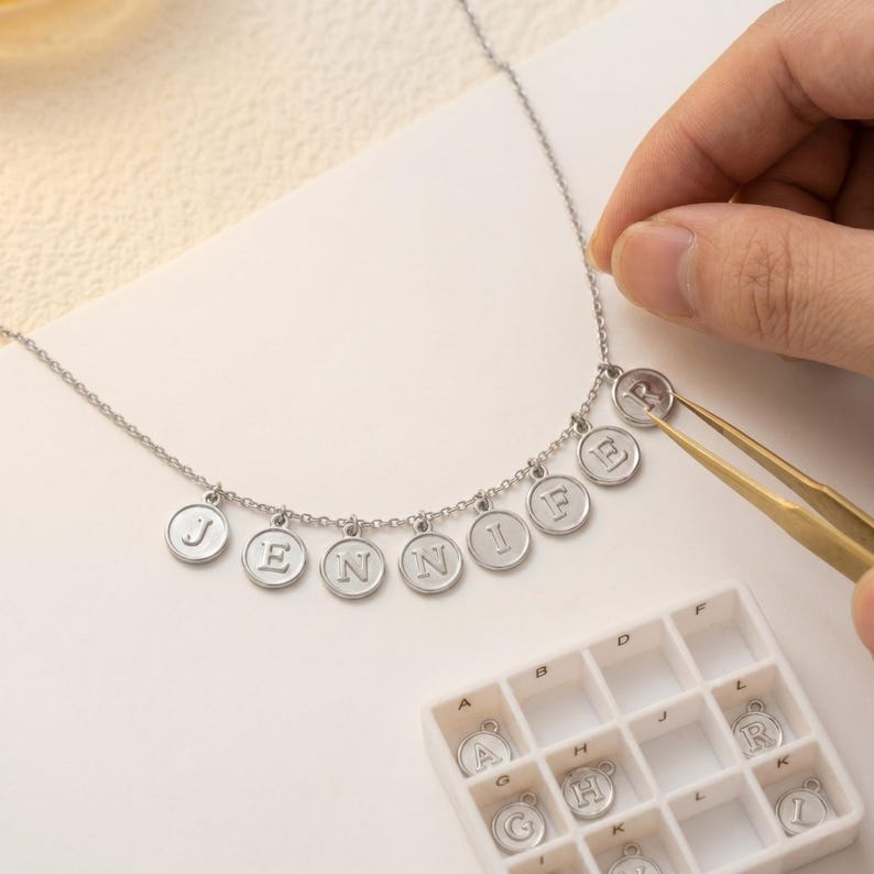 May include: A silver necklace spelling out "JENNI" with individual letter charms. The letter "R" is being added with tweezers. A small tray with spare letter charms is in the foreground.