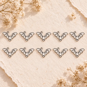May include: Ten identical silver-toned metal bat charms arranged in two rows of five. Each charm features a stylized bat with outstretched wings and a small circular loop at the top for attachment, showcasing a detailed embossed design.