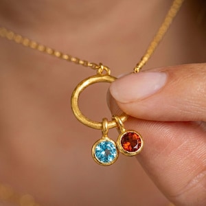 May include: A gold-colored necklace with a circular pendant and two gemstone charms. One charm is a light blue gemstone, and the other is a red-orange gemstone. The necklace is held by a person.