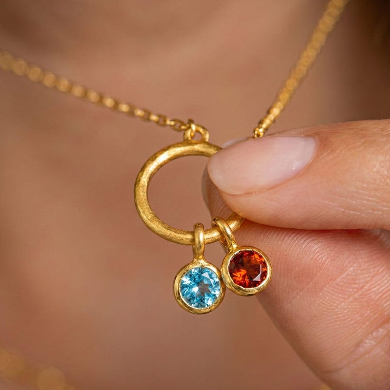 May include: A gold-colored necklace with a circular pendant and two gemstone charms. One charm is a light blue gemstone, and the other is a red-orange gemstone. The necklace is held by a person.