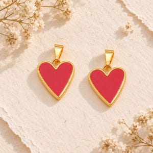 May include: Two heart-shaped pendants featuring a vibrant red enamel center encased in a polished gold-tone metal frame. Each pendant includes a small gold-tone bail at the top, designed for attachment to a necklace chain or other jewelry component.