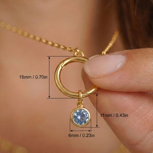 May include: A gold-colored necklace with a circular pendant and a small, round, light blue gemstone. The pendant is 0.70 inches in diameter, and the gemstone is 0.23 inches in diameter. The necklace is being held by a person.