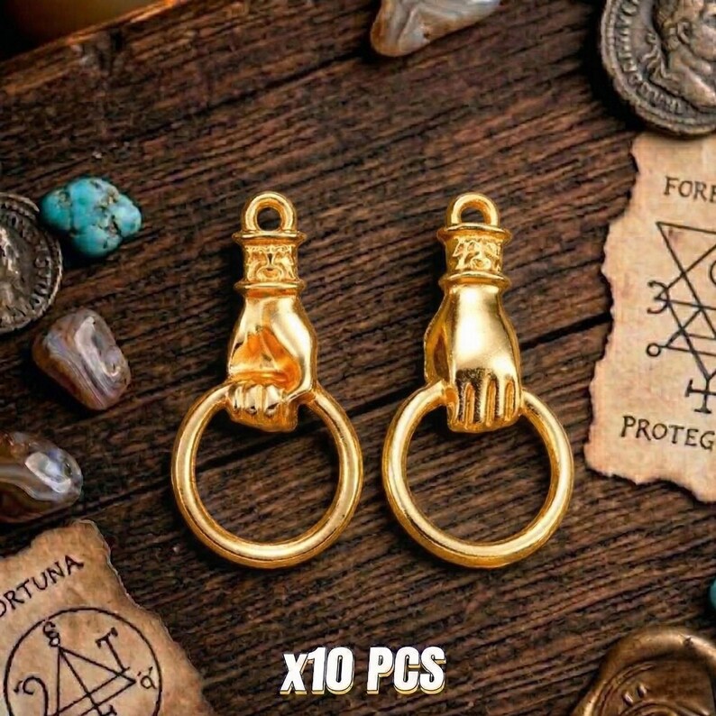 May include: Two gold-colored pendants shaped like a hand holding a ring. The pendants have a loop at the top for hanging. The text "X10 PCS" is at the bottom. The pendants are on a wooden surface with other decorative items.