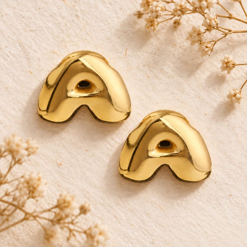 May include: A pair of gold-toned stud earrings shaped like the letter A. The earrings feature a smooth, polished, and reflective metallic surface with a rounded, puffed design. Each letter has a small, dark, recessed center point.