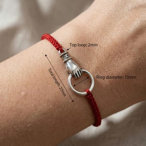 May include: Red cord bracelet featuring a silver hand clasping a silver ring. The total length is 2.7cm, the ring diameter is 1.3cm, and the top loop is 2mm.
