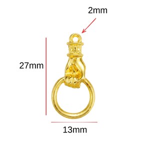 Puede incluir: Un dije dorado con forma de mano que sostiene un anillo. El dije mide 27 mm de largo, 13 mm de ancho y tiene un lazo de 2 mm. Probablemente est&aacute; hecho de metal y est&aacute; dise&ntilde;ado para la fabricaci&oacute;n de joyas o manualidades.