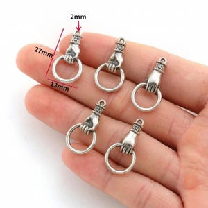 May include: Silver-coloured metal charms shaped like a hand holding a ring. Each charm is approximately 2.7cm long, 1.3cm wide, and 2mm thick. The charms are displayed on a light-coloured surface.