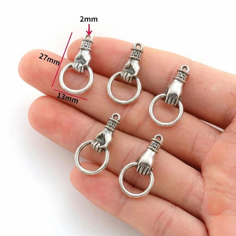 May include: Silver-coloured metal charms shaped like a hand holding a ring. Each charm is approximately 2.7cm long, 1.3cm wide, and 2mm thick. The charms are displayed on a light-coloured surface.