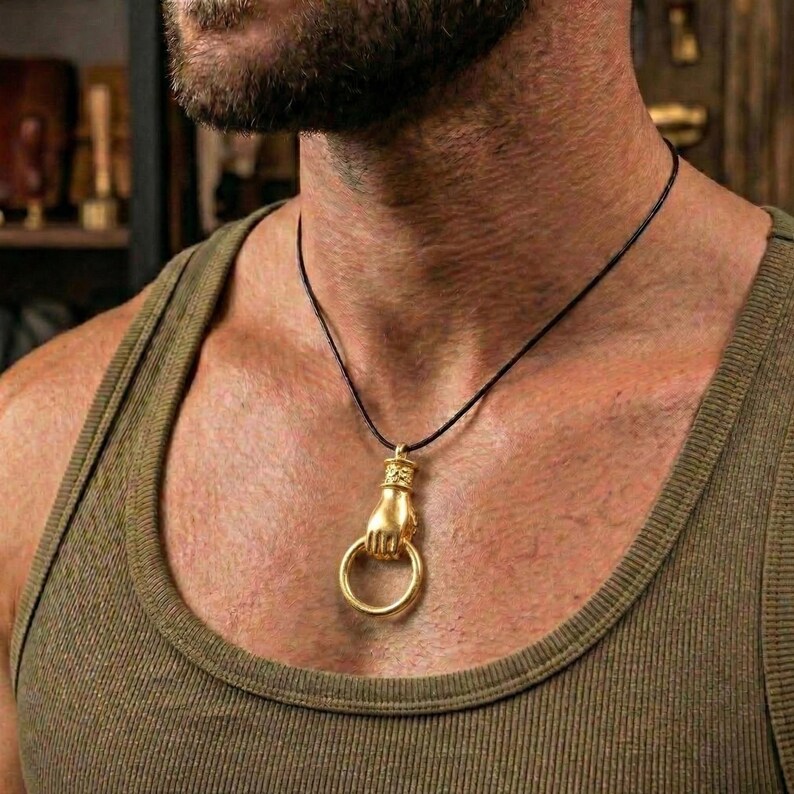 May include: Gold-colored pendant necklace featuring a hand holding a ring. The pendant is suspended from a black cord. The necklace is worn against a green tank top.