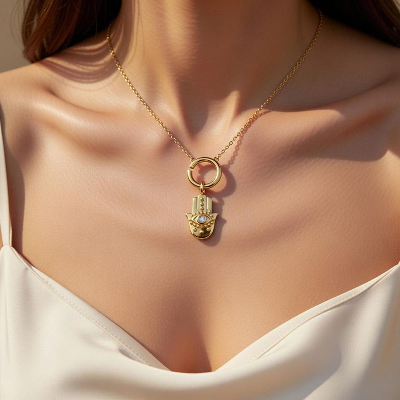 May include: Gold-toned necklace featuring a Hamsa hand pendant with an eye design and a small, round, white gemstone. The pendant hangs from a delicate chain. The necklace is worn against a white top.