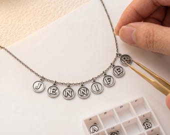 May include: A silver-toned necklace with the name "JENNIF" spelled out in individual letter charms. The letters are in a classic font and are suspended from a delicate chain. A person is using tweezers to add the letter "R" to the necklace.