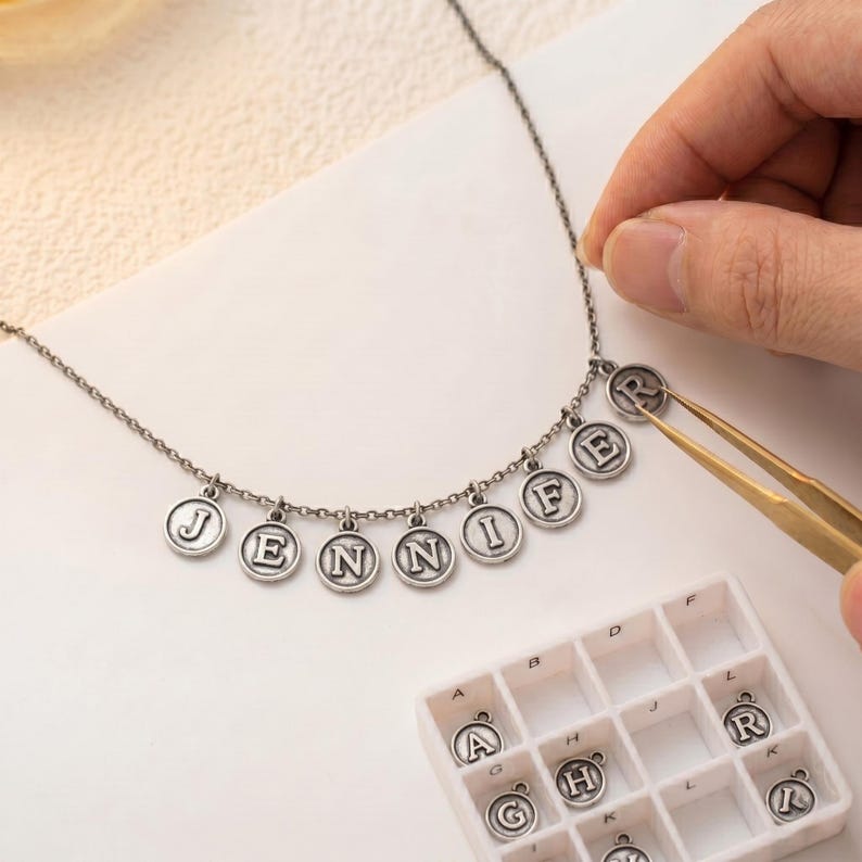 May include: A silver necklace spelling "JENNIF" with individual letter charms. The letters are in a classic font and hang from a delicate chain. A person is using tweezers to add the letter "R" to the necklace.