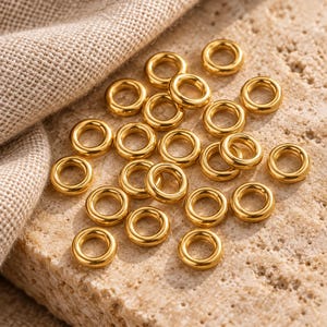 Gold-tone stainless steel closed jump rings. Textured hero view. 4mm outer diameter; 1.2mm wire. Buyer pack: 50 pieces.