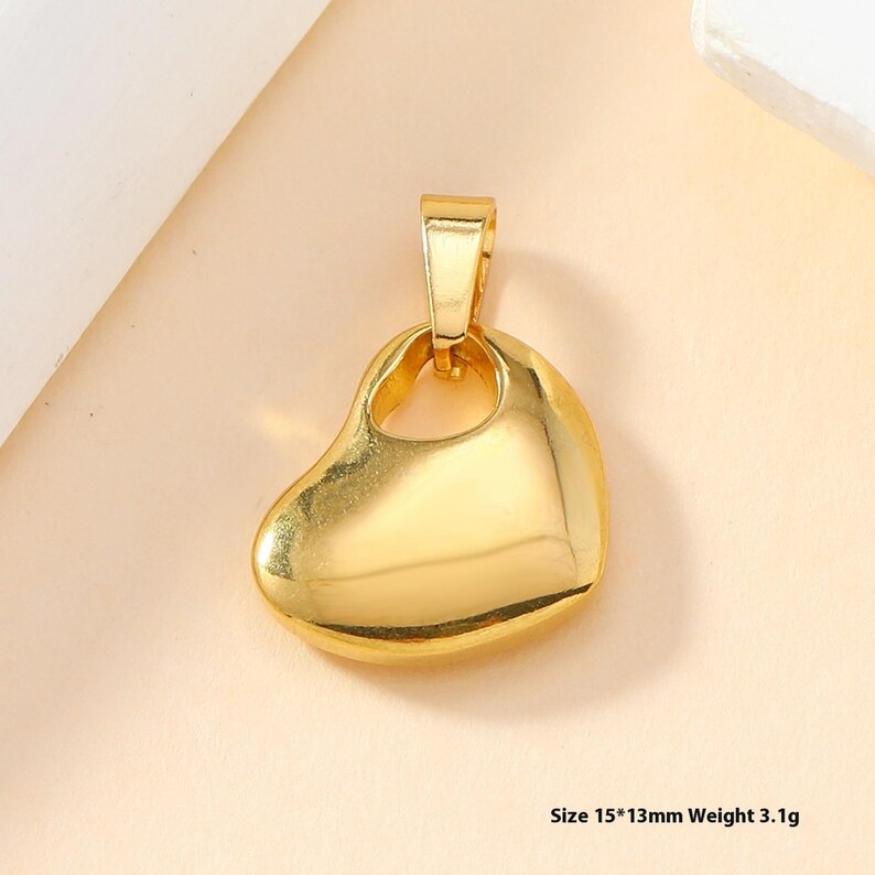 May include: A gold-toned metal pendant shaped like a smooth, asymmetrical heart with a bail at the top. The pendant has a polished, reflective surface and measures 15 by 13 millimeters with a weight of 3.1 grams as indicated in the image text.