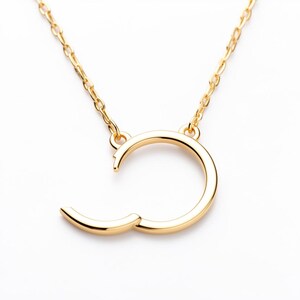 May include: A gold-colored necklace with a unique open-circle pendant. The pendant has a curved design with a small, hinged section that opens. The necklace chain is also gold-colored and delicate, creating a minimalist jewelry piece.