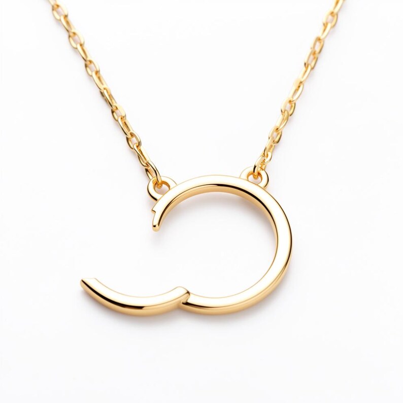 May include: A gold-colored necklace with a unique open-circle pendant. The pendant has a curved design with a small, hinged section that opens. The necklace chain is also gold-colored and delicate, creating a minimalist jewelry piece.