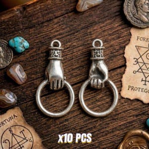 May include: Two silver-coloured hand-shaped charms holding a ring, with a loop at the top. The charms are displayed on a wooden surface, with the text "X10 PCS" in yellow. Additional decorative elements include coins and gemstones.