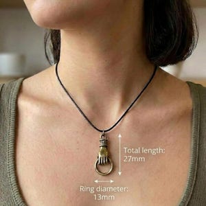 May include: A bronze-colored pendant necklace featuring a hand holding a ring. The total length is 27mm, and the ring diameter is 13mm. The necklace has a black cord. The pendant is a unique accessory.