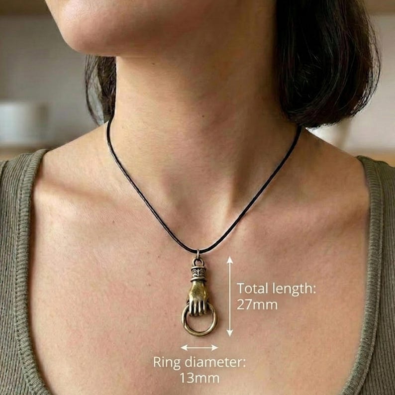 May include: A bronze-colored pendant necklace featuring a hand holding a ring. The total length is 27mm, and the ring diameter is 13mm. The necklace has a black cord. The pendant is a unique accessory.