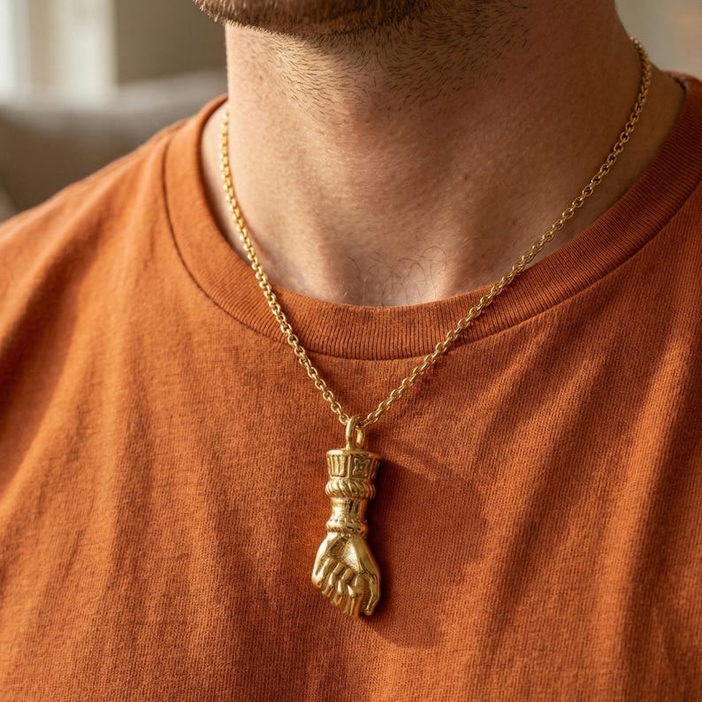May include: Gold-colored pendant necklace featuring a detailed hand holding a fist. The pendant hangs from a delicate chain. The person in the image is wearing an orange t-shirt. The necklace is a piece of jewelry.