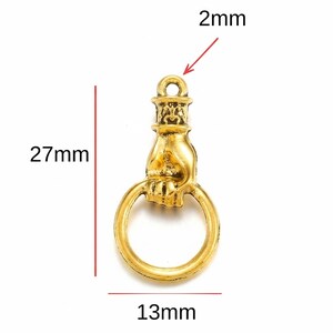 May include: A gold-colored charm shaped like a hand holding a ring. The charm is 27mm long, 13mm wide, and has a 2mm hole at the top. The charm has an antique or vintage aesthetic.