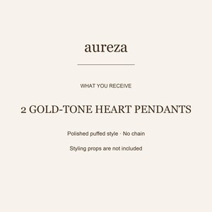 May include: A product information card with the text aureza, WHAT YOU RECEIVE, 2 GOLD-TONE HEART PENDANTS, Polished puffed style, No chain, and Styling props are not included. The text is dark brown on a light cream background with a horizontal line.