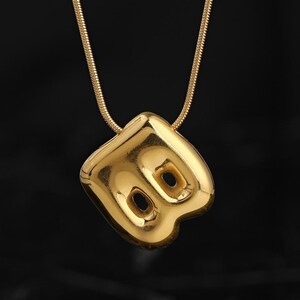 May include: A gold-toned necklace featuring a polished, puffed pendant in the shape of the letter B. The pendant has a smooth, reflective surface with two distinct circular cutouts within the letter's structure, suspended from a thin snake chain.