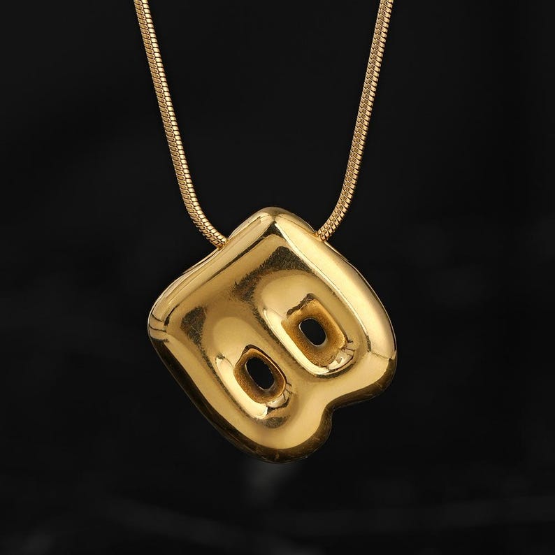 May include: A gold-toned necklace featuring a polished, puffed pendant in the shape of the letter B. The pendant has a smooth, reflective surface with two distinct circular cutouts within the letter's structure, suspended from a thin snake chain.