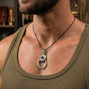 May include: Silver pendant necklace with a hand grasping a ring. The pendant is suspended from a thin, black cord. The necklace is worn against a person's chest, with a green vest top visible.