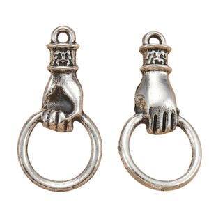 May include: Two silver-toned metal charms, each shaped like a hand grasping a circular ring. The hand features a cuff detail at the wrist and a loop at the top for attachment. The metal has a weathered, antiqued finish with dark recessed areas.